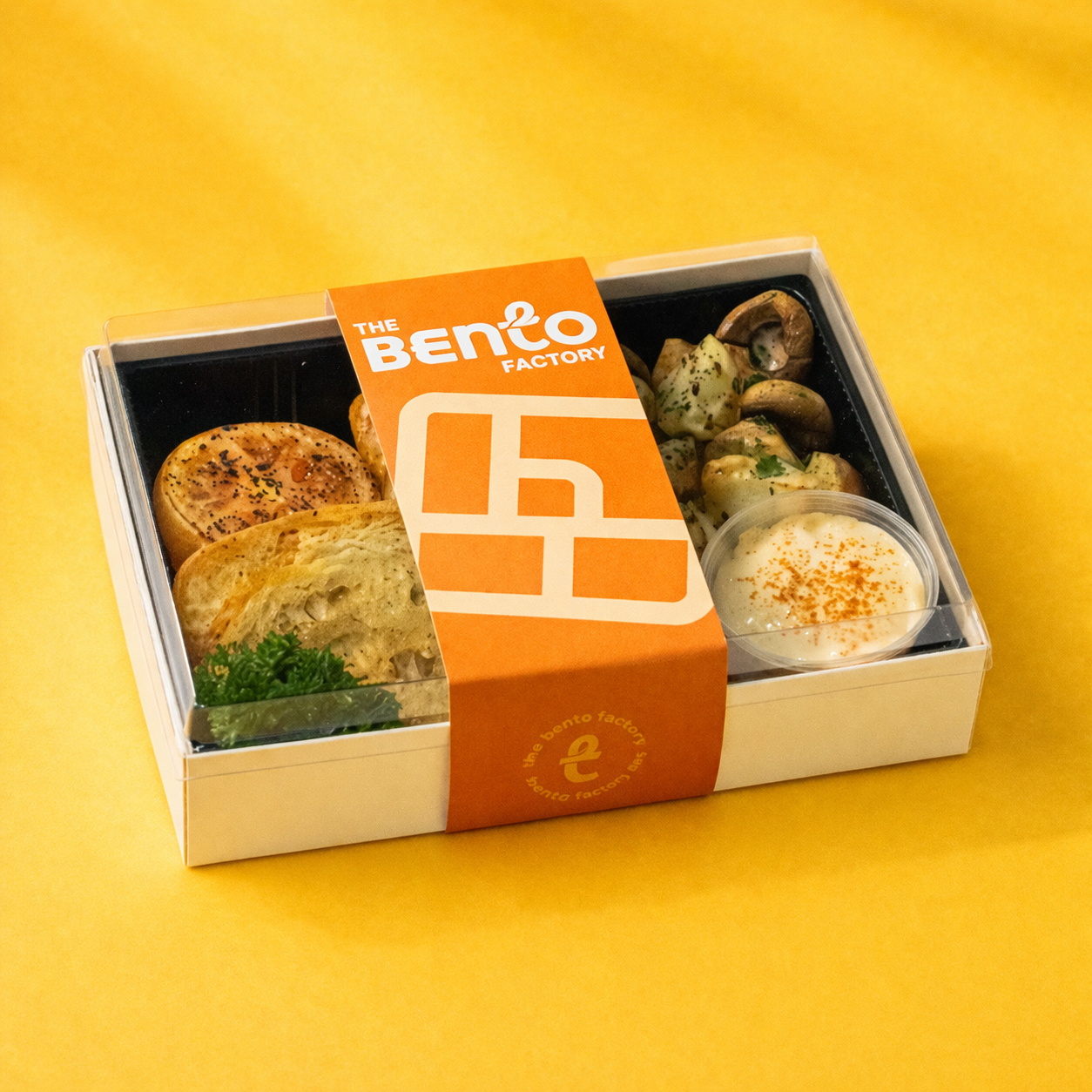 Bento box with food items on a yellow background, featuring 'The Bento Factory' branding.