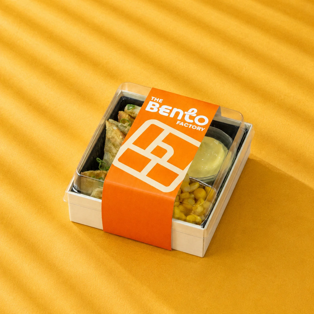 Bento box with food items on a yellow background, featuring 'The Bento Factory' branding.