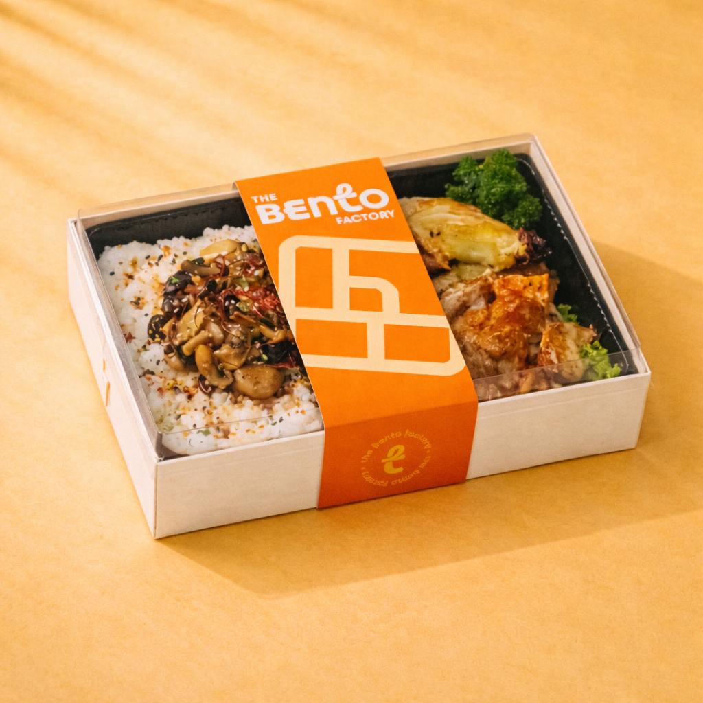 Bento box with sautéed mushrooms over rice, roasted chicken, and greens, packaged in a cream box with an orange “The Bento Factory” sleeve on a warm yellow gradient background.